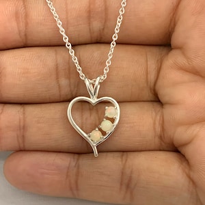 May include: A silver-toned necklace featuring a heart-shaped pendant. The pendant has three small, round, opalescent stones set within the heart's outline. The chain is delicate and appears to be made of silver.