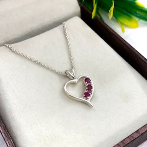 Op de afbeelding: Een zilveren ketting met een hartvormige hanger met vier rode robijnstenen die in het hart zijn gezet. De ketting ligt op een witte achtergrond.