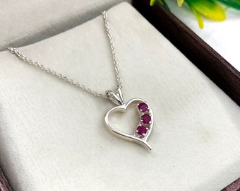 May include: A silver heart-shaped pendant necklace with four red ruby stones set in the heart. The necklace is on a white background.