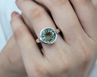 May include: A silver ring featuring a large, round, light green gemstone. The gemstone is set in a bezel setting, and the ring is shown on a person's finger. The ring is a piece of jewelry.