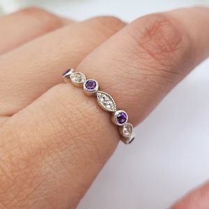 Puede incluir: Un anillo de banda de plata con una fila de pequeñas piedras preciosas púrpuras y piedras de circonita cúbica transparentes.