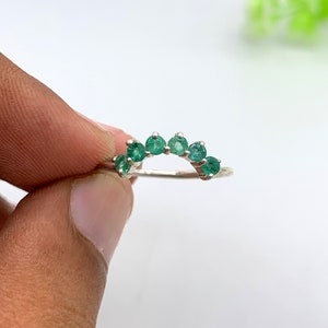 Op de afbeelding: Een zilveren ring met een gebogen rij van zeven kleine, ronde, smaragdgroene edelstenen. De ring wordt tegen een witte achtergrond gehouden, waardoor de levendige kleur van de edelstenen en het delicate ontwerp van de ring worden benadrukt.
