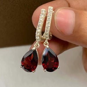 May include: A pair of silver drop earrings featuring teardrop-shaped deep red garnet stones suspended from hoops encrusted with small clear white stones. The metal has a polished finish, and the design showcases a faceted cut on the red gems.