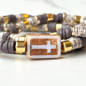 May include: Three layered bracelets with gray, white, and gold beads. The top bracelet has a square charm with a white cross on a brown background.