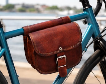 May include: A brown leather bicycle frame bag with a flap closure, secured by a silver-toned metal buckle. The bag is attached to the top tube of a metallic blue bicycle frame, featuring visible stitching along the edges and a structured design.