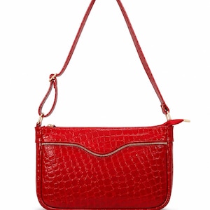May include: A red handbag with a crocodile-effect texture. The bag features a long shoulder strap and a gold zip. The rectangular bag has rounded corners. The zip is curved. The bag is made of a shiny material.