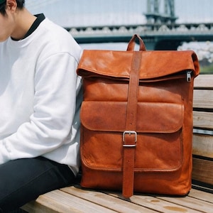 May include: A tan leather roll-top backpack featuring a large front pocket with a vertical strap and a silver metal buckle closure. The bag has a rectangular shape with visible stitching along the edges and a top handle. The text BAG PACKS is visible.