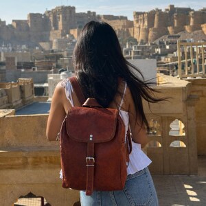 May include: A brown leather backpack with a flap and buckle closure. The person is standing on a rooftop overlooking a city with beige buildings under a blue sky. The backpack is a classic style, suitable for everyday use.