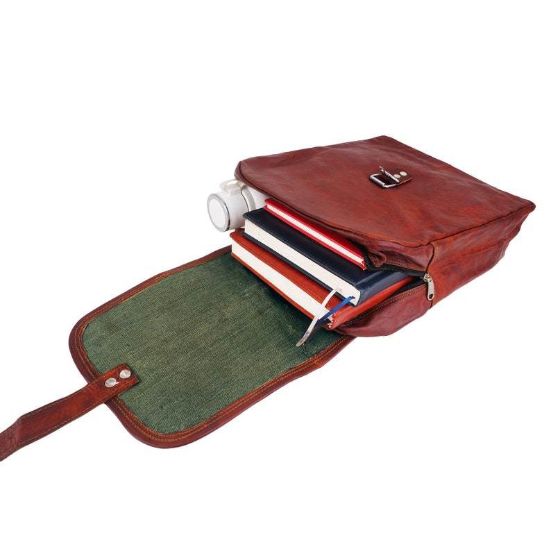 May include: A brown leather satchel with a flap, open to reveal a green interior and several books. A white cylindrical object is also inside. The bag has a metal clasp and a zipper closure.