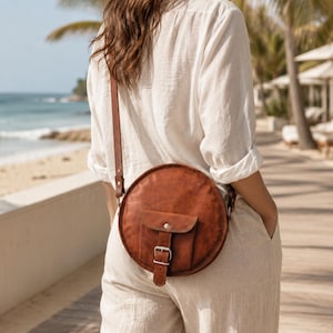 May include: A round, brown leather crossbody bag featuring a front flap pocket with a silver-toned metal buckle closure and a matching adjustable shoulder strap. The bag has a smooth, consistent texture and a circular silhouette.