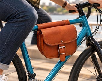 May include: A tan leather bicycle frame bag featuring a rounded flap, visible contrast stitching, and a silver-toned metal buckle closure. The bag is securely mounted to the top tube of a bright blue bicycle frame, providing storage space.