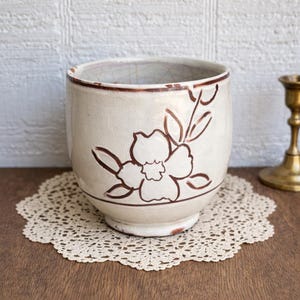 May include: Cream-colored ceramic planter with a brown floral design. The planter has a wide, rounded shape and a slightly distressed appearance. The flower design is hand-drawn and adds a touch of rustic charm. The planter sits on a lace doily.