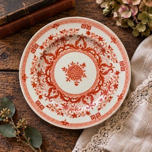 May include: A round ceramic plate featuring a white base with a detailed orange floral pattern. The design includes a central flower motif surrounded by intricate scrolling vines and geometric border accents along the outer rim.