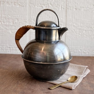 May include: A vintage silver-toned metal pitcher with a woven handle and lid. The pitcher has a rounded body and a spout. A small gold-colored spoon rests on a linen cloth next to the pitcher. The pitcher is likely used for serving beverages.