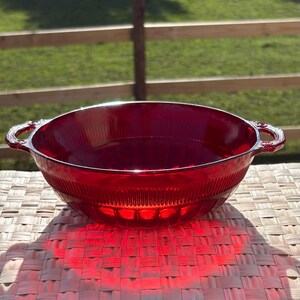 May include: A translucent ruby red glass bowl featuring a circular shape with two integrated side handles. The exterior surface of the bowl displays a vertical ribbed pattern around the upper rim and a series of rounded indentations along the base.