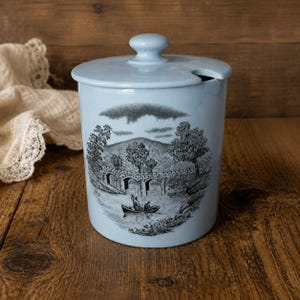May include: A light blue ceramic canister with a matching lid featuring a small notch. The exterior displays a detailed black transferware illustration of a stone bridge over a river with two people in a small boat and trees in the background.