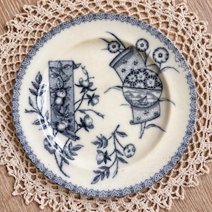 May include: A round ceramic plate with an off-white base and dark blue transferware patterns. The rim features a geometric lace-like border. The center displays two rectangular scrolls, one with fruit and one with a circular landscape scene of a boat.