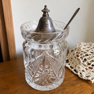 May include: A clear cut glass jar featuring a geometric starburst pattern and a scalloped rim. The jar includes a removable silver-toned metal lid with a small finial top and a matching silver-toned metal spoon resting inside the container.