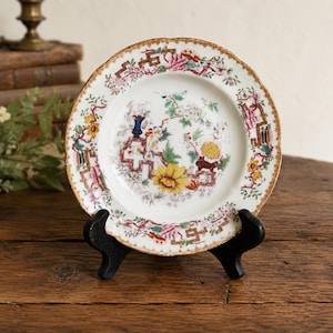 May include: A round ceramic plate featuring a white base with a colorful floral pattern, including yellow, pink, and green blossoms. The rim is decorated with a gold-toned border and geometric lines, resting on a black display stand.