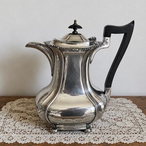 May include: A silver-toned metal teapot featuring a curved spout, a hinged lid with a dark finial, and a prominent black angular handle. The body has a multi-faceted, bulbous shape with vertical panels and rests on small, rounded feet.
