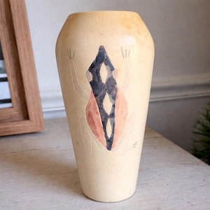 May include: A beige ceramic vase with a hand-painted design. The design features a central diamond shape in black and red, with a stylized frog outline. The vase has a tall, cylindrical shape and a slightly flared top.