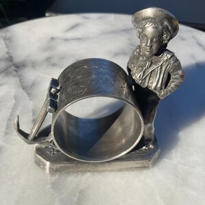 May include: A silver-toned napkin ring featuring a sculpted figure of a young boy in a sailor hat and jacket. The ring is cylindrical with floral detailing and a hinged arm. The boy figure is standing on a rectangular base.