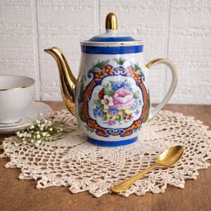 May include: A white and blue porcelain teapot with gold accents, featuring a floral design. The teapot sits on a cream-colored doily, with a gold spoon nearby. A white teacup with a gold rim is also visible.