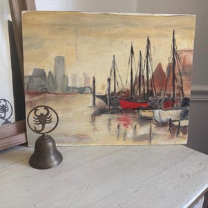 May include: A rectangular oil painting on canvas depicting a harbor scene with sailboats, a red boat, and a distant city skyline in shades of beige and brown, accompanied by a brass bell featuring a scorpion emblem on its handle.