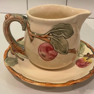May include: Cream-colored ceramic pitcher and saucer set. The pitcher has a curved handle and a spout. The set is decorated with a branch design featuring red apples and green leaves. The saucer has a scalloped edge.