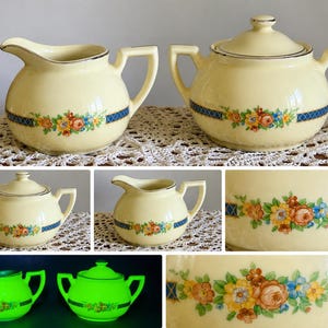 May include: A cream-colored ceramic creamer and sugar bowl set with a lid, featuring a floral pattern of orange, yellow, and blue flowers accented by a blue cross-hatch border and silver trim. The set glows bright green under ultraviolet light.