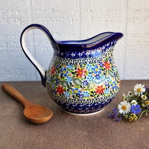 May include: A ceramic pitcher with a blue rim and handle, adorned with a floral pattern in red, blue, and yellow. The pitcher has a curved spout and a white interior. A wooden spoon rests beside it.
