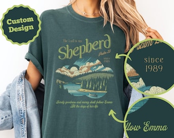 Może przedstawiać: Ciemnozielona koszulka z napisem "The Lord is my Shepherd" i krajobrazem. Projekt obejmuje góry, jezioro i drzewa. Koszulka ma również napis "Since 1989" i "Follow Emma".