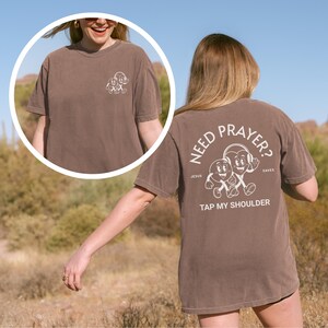 May include: Brown t-shirt with white graphic on front and back. The front graphic is a cartoon design. The back of the shirt says "Need Prayer? Tap My Shoulder" with a cartoon design. The shirt is worn by a person.