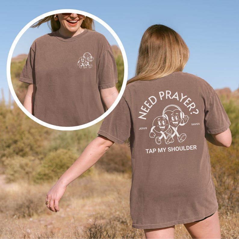 May include: Brown t-shirt with white graphic on front and back. The front graphic is a cartoon design. The back of the shirt says "Need Prayer? Tap My Shoulder" with a cartoon design. The shirt is worn by a person.