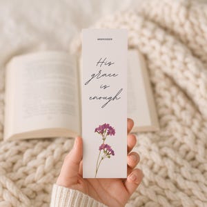 May include: A white bookmark with the text "His grace is enough" in cursive script, and the hashtag #REMINDER at the top. The bookmark features a sprig of purple flowers at the bottom. The background includes an open book and a cream-colored knitted blanket.