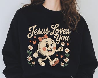 Może przedstawiać: Czarna bluza z napisem "Jesus Loves You" w stylu vintage. Grafika przedstawia uśmiechniętą postać z kreskówki z kokardą, otoczoną kwiatami i sercami. Swobodny, wygodny styl.