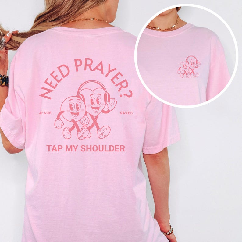 May include: A light pink t-shirt with a graphic on the back featuring two cartoon characters and the text "NEED PRAYER? TAP MY SHOULDER." The design is in a matching pink color. A smaller version of the graphic is on the front left chest.