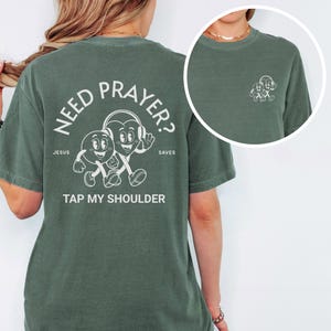 May include: Green t-shirt with white text and graphics. The back of the shirt says "NEED PRAYER?" with cartoon heart characters and the words "JESUS SAVES TAP MY SHOULDER." A smaller graphic is on the front. The shirt is made of a soft material.
