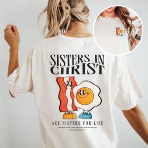 Puede incluir: Camiseta color crema con el texto "SISTERS IN CHRIST" sobre una tira de tocino de dibujos animados y un huevo frito. Debajo, se lee "ARE SISTERS FOR LIFE" con un versículo bíblico. Un gráfico más pequeño del huevo y el tocino está en el pecho izquierdo.
