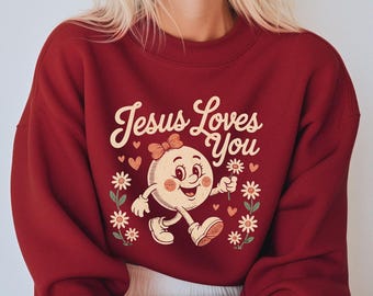 Może przedstawiać: Bordowa bluza z napisem "Jesus Loves You" w stylu vintage. Grafika przedstawia uśmiechniętą, kreskówkową postać trzymającą kwiat, otoczoną stokrotkami i sercami.