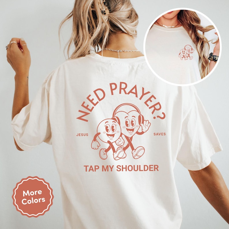 May include: Cream-colored t-shirt with a retro-style graphic in a reddish-brown hue. The graphic features two cartoon characters, one a heart wearing headphones, and the other a round character. Text on the shirt reads "NEED PRAYER? TAP MY SHOULDER" and "JESUS SAVES".