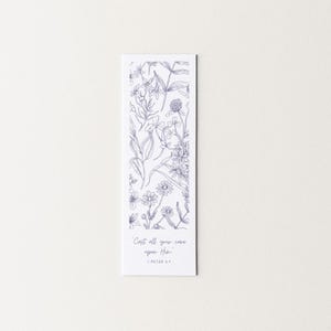 May include: A rectangular white bookmark with a detailed blue floral illustration. The design includes various flowers and foliage. The text "Cast all your care upon Him" and "1 Peter 5:7" are printed in a delicate script at the bottom.