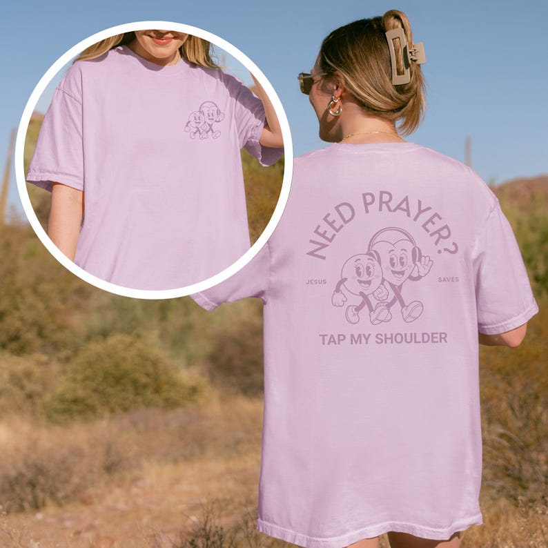 May include: A light purple t-shirt with a cartoon graphic on the front and back. The front graphic is a small cartoon character. The back graphic says "NEED PRAYER? TAP MY SHOULDER" with cartoon characters. The shirt is a casual style.