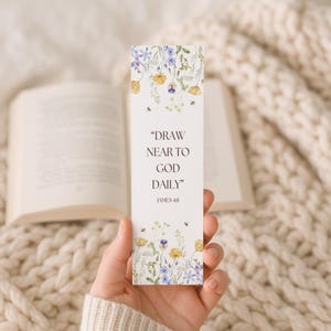 May include: A white bookmark with floral illustrations in yellow, blue, and green, held in a hand. The bookmark features the text "DRAW NEAR TO GOD DAILY" and "JAMES 48". An open book and a knitted blanket are in the background.