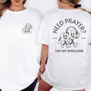 May include: White t-shirt with a black and white graphic on the front and back. The front features a small cartoon design. The back has the text "NEED PRAYER?" with a cartoon heart and circle character design, and the text "TAP MY SHOULDER."