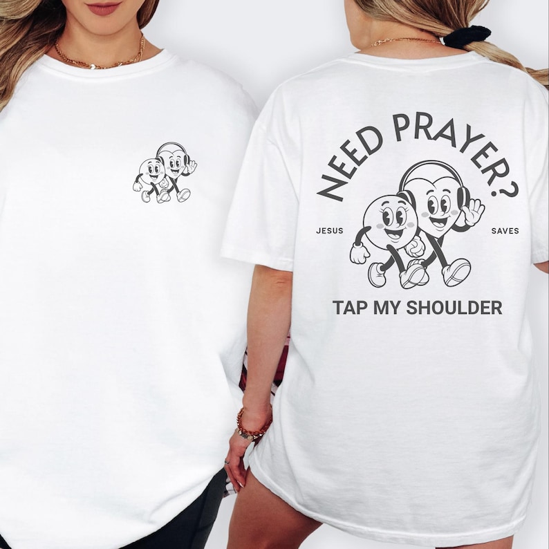 May include: White t-shirt with a black and white graphic on the front and back. The front features a small cartoon design. The back has the text "NEED PRAYER?" with a cartoon heart and circle character design, and the text "TAP MY SHOULDER."