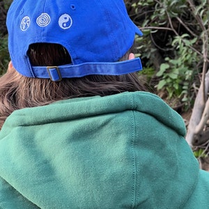 May include: A blue baseball cap with a white embroidered design of a globe, a spiral, and a yin yang symbol. The cap is worn by a person wearing a green hooded sweatshirt with a white circle design on the back.