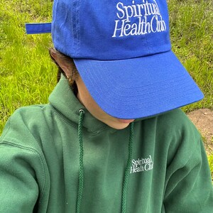 May include: A person wearing a green hoodie with the text "Spiritual Health Club" embroidered on the front. They are also wearing a blue baseball cap with the same text embroidered on the front.