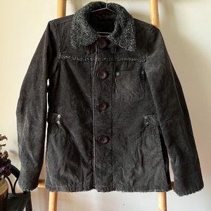 Puede incluir: Una chaqueta de pana gris oscuro con un cuello de pelo gris y detalles de costura roja. La chaqueta tiene cierre de botones y un bolsillo de parche en el pecho izquierdo.