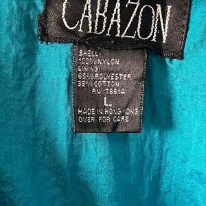 Pode incluir: Uma jaqueta azul turquesa com uma etiqueta preta que diz "CABAZON" e lista o conte&uacute;do do tecido como 100% nylon, 65% poli&eacute;ster e 35% algod&atilde;o. A etiqueta tamb&eacute;m diz "Feito em Hong Kong" e "Virar para o cuidado".